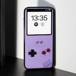 Puede incluir: Una funda de teléfono lila diseñada para parecerse a una consola de juegos portátil retro. La funda presenta una cruceta negra, botones rojos y los textos "SELECT" y "START". La pantalla del teléfono muestra la hora 13:35.
