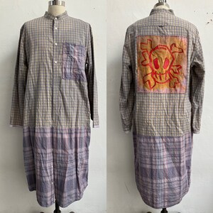 May include: A long-sleeved, plaid shirt with a skull and crossbones design on the back. The shirt is made of a light blue and beige plaid fabric and has a patch pocket on the left chest. The bottom half of the shirt is a different plaid pattern in shades of purple, pink, and blue.