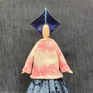 May include: A wooden doll wearing a pink and white tie-dye sweater, a blue floral skirt, and a blue and black hat with a beaded trim.