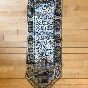 May include: A tapestry wall hanging with a gold and black border and a white background. The tapestry features Arabic calligraphy and a depiction of the Kaaba, a cube-shaped building in Mecca, Saudi Arabia. The tapestry is a religious artifact and is often used in Islamic homes and places of worship.