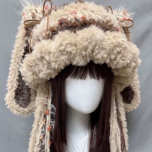 Puede incluir: Un gorro de punto esponjoso marrón y blanco con largos hilos de lana colgantes. El gorro tiene orejeras y un pompón en la parte superior.