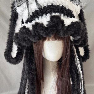 May include: Black and white crocheted bunny hat with long fur ear and paw accents. The hat is on a mannequin with long brown hair.