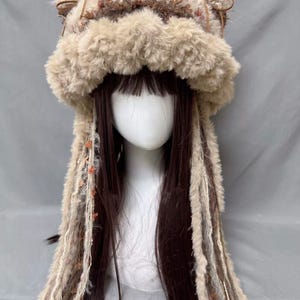 May include: A fluffy brown and white knit hat with long, dangling strands of yarn. The hat has cat ears and a furry brim.