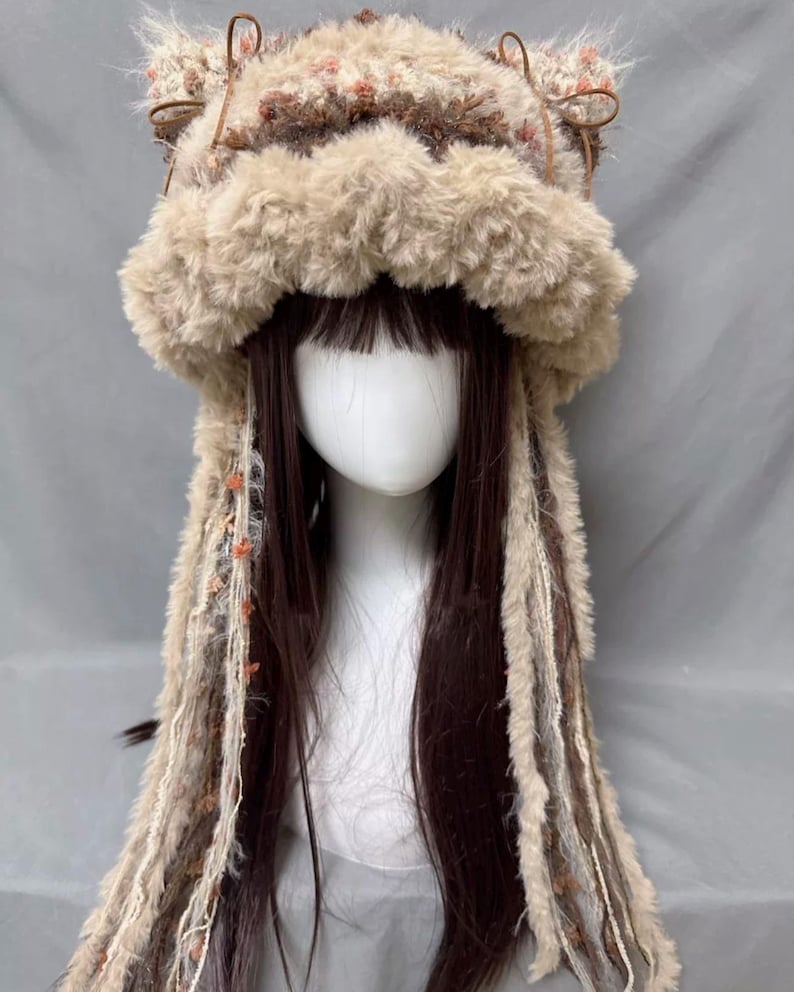 May include: A fluffy brown and white knit hat with long, dangling strands of yarn. The hat has cat ears and a furry brim.
