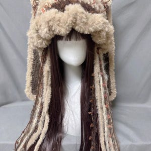 May include: A brown and beige knitted hat with furry ear flaps and long fringe. The hat has a crocheted design and is decorated with small brown beads.