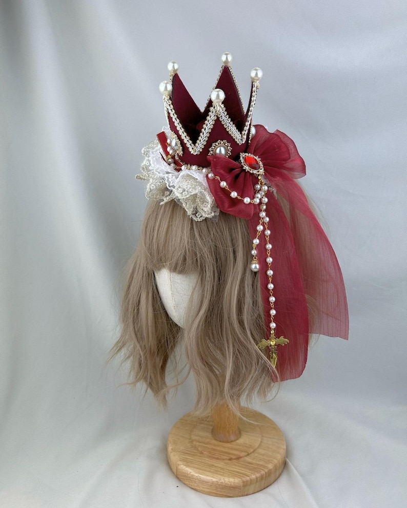 Puede incluir: Una corona de terciopelo burdeos con ribetes dorados y detalles de perlas. La corona est&aacute; adornada con un lazo rojo y una cadena de perlas y oro con un colgante de cruz. La corona est&aacute; sobre una cabeza de maniqu&iacute; con cabello casta&ntilde;o.