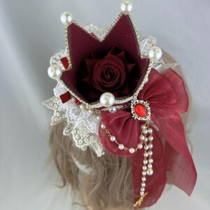 Puede incluir: Una corona burdeos con una rosa roja en su interior, adornada con perlas blancas y un lazo rojo con una joya roja en el centro. La corona est&aacute; sobre una cabeza de maniqu&iacute; con cabello casta&ntilde;o.