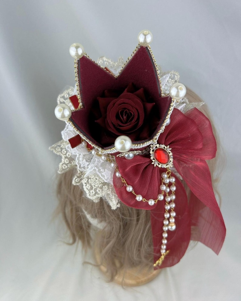 Puede incluir: Una corona burdeos con una rosa roja en su interior, adornada con perlas blancas y un lazo rojo con una joya roja en el centro. La corona est&aacute; sobre una cabeza de maniqu&iacute; con cabello casta&ntilde;o.