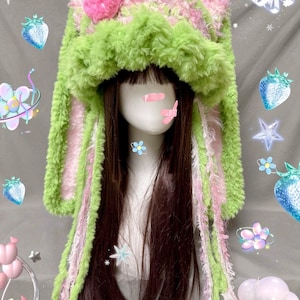 May include: Green and pink fuzzy bunny ear hat with a strawberry on top. The hat is worn on a mannequin with long brown hair.