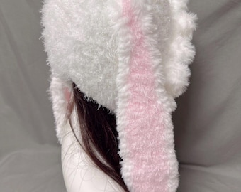 Könnte beinhalten: Weißer, flauschiger Hut mit langen, rosa Hasenohren. Der Hut ist aus weichem, flauschigem Material und hat ein schlichtes Design.