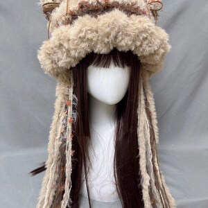 May include: A brown and beige fuzzy hat with long, brown, beige, and white yarn strands hanging down. The hat has two furry ear flaps on top.