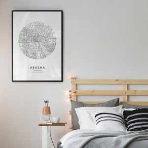 May include: A black framed print of a map of Arusha, Tanzania, hanging on a white wall above a bed with a wooden headboard and gray bedding. The bed has a white pillow, a gray pillow, and a black and white striped pillow.