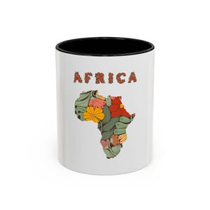 May include: White ceramic mug with a black interior. The mug features a colorful floral design in the shape of the African continent, with the word "Africa" above it in a floral font. The design includes red, yellow, and green flowers.