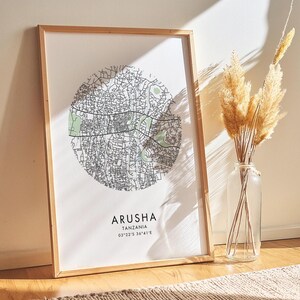May include: A framed print of a map of Arusha, Tanzania. The map is in a circle and shows the city streets. The print is on a white background with the city name and coordinates printed below the map.