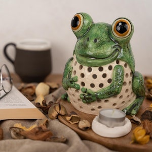 Peut inclure: Un bougeoir en céramique en forme de grenouille avec un glaçage vert et blanc. La grenouille a de grands yeux et un large sourire. Le bougeoir a un trou au centre pour une bougie chauffe-plat.