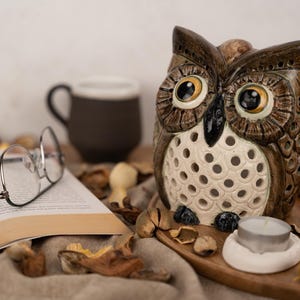 May include: A ceramic owl candle holder with a white and brown design. The owl has large, round eyes and a detailed beak. The candle holder is sitting on a wooden tray with a tea light candle inside.