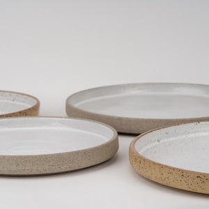 Puede incluir: Un juego de tres platos redondos de cerámica con un exterior beige moteado y una superficie interior blanca, lisa y brillante. Cada plato tiene un borde bajo elevado y una base plana, mostrando una textura natural y terrosa.