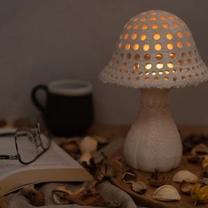 May include: A white ceramic mushroom lamp with a light inside, casting a warm glow. The lamp is on a table with a book, glasses, and dried flower petals.