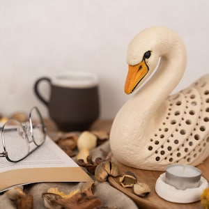 May include: A white ceramic swan candle holder with a decorative cutout design. The swan has a yellow beak and black eyes. The candle holder is sitting on a wooden surface with a small white tea light candle inside.