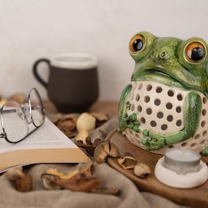 May include: A ceramic frog candle holder with a white, perforated body and green accents. The frog has large, round eyes and a wide mouth. The candle holder is sitting on a wooden surface with a tea light candle inside.