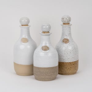 Puede incluir: Tres botellas de aceite de cerámica con tapones esféricos. Cada botella tiene una etiqueta redonda con la palabra OIL en relieve. La botella de la izquierda es blanca lisa y beige, la central es blanca moteada y beige, y la derecha es blanca moteada y marrón.