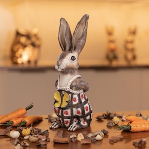 Op de afbeelding: Een bruin konijnenbeeldje dat een rood-wit geruit jurkje draagt met gele vlinderaccenten. Het beeldje staat op een houten oppervlak met verspreide bruine en witte decoratieve items.