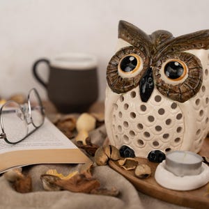 May include: A white ceramic owl candle holder with brown and black details. The owl has large, round eyes and a detailed beak. The candle holder has a pattern of small holes throughout the body. A small white candle is sitting inside the owl's body.