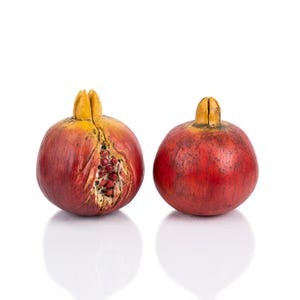Op de afbeelding: Twee rode granaatappels met gele toppen, een met een spleet die de zaden erin laat zien. De granaatappels staan op een witte achtergrond.