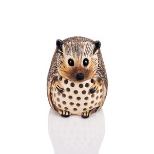 May include: A ceramic hedgehog figurine with a brown and white patterned body. The hedgehog is curled up with its head and legs tucked in, and its eyes are closed. The figurine has a textured surface and a hole in the center of its body.