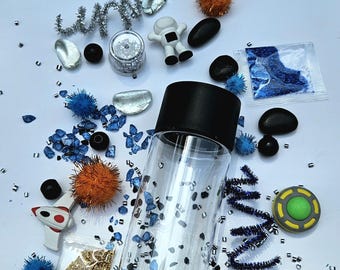 Puede incluir: Una botella sensorial de plástico transparente con tapa negra, llena de pequeñas piedras negras y azules. Alrededor de la botella hay artículos con temática espacial, incluyendo un astronauta, un cohete de juguete, un OVNI y pompones.