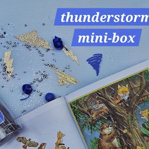 May include: A "thunderstorm mini-box" with glitter, blue accents, and a children's book illustration of foxes in a tree. The box includes gold glitter lightning bolts and cloud shapes.
