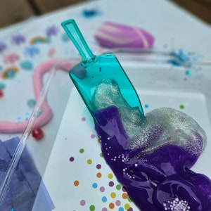 Może przedstawiać: Fioletowy i srebrny slime z brokatem, który jest nabierany z turkusowej plastikowej łopatki na białą powierzchnię z kolorowymi konfetti.