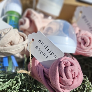 May include: A gift basket with baby bottles, rolled pink and beige fabric items, and a tag reading "phillips avent." The basket is filled with green shredded paper. The bottles are clear with blue and green accents.