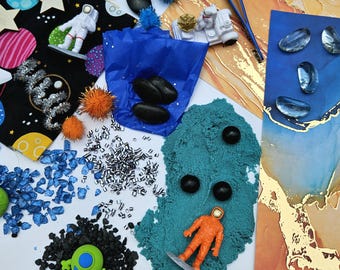 May include: A colorful, whimsical arrangement of space-themed toys and craft supplies. Includes astronaut figurines, a toy space rover, blue and orange pom-poms, black stones, blue and silver glitter, and a green alien figure. The background features abstract art.