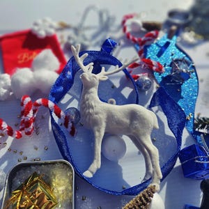Könnte beinhalten: Ein weißes Rentier schmückt eine Weihnachtsdekoration, umgeben von einem blauen Band. Rot-weiß gestreifte Zuckerstangen, eine kleine rote Tasche und eine blaue Glitzerverzierung ergänzen die Szene. Eine kleine Dose mit einem goldenen Geschenk ist ebenfalls vorhanden.