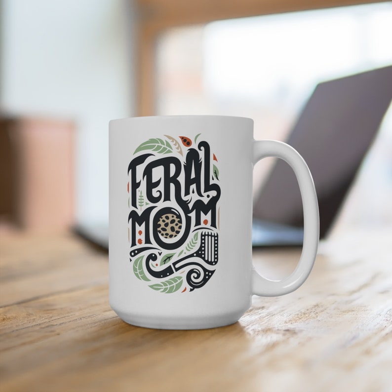 Pu&ograve; includere: Tazza da caff&egrave; in ceramica bianca con un design nero e verde che dice "Feral Mom" in un font stilizzato.