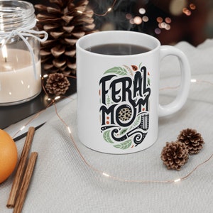 Pu&ograve; includere: Una tazza da caff&egrave; in ceramica bianca con un design nero e verde che dice "Feral Mom". La tazza &egrave; piena di caff&egrave; e c'&egrave; del vapore che esce da essa.