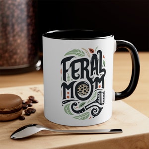 Pu&ograve; includere: Tazza in ceramica bianca con bordo e manico neri. La tazza presenta un'illustrazione in nero e verde di una "Feral Mom" stilizzata con un motivo leopardato.