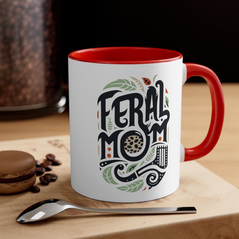Pu&ograve; includere: Tazza in ceramica bianca con bordo e manico rossi. La tazza presenta un'illustrazione in nero e verde di un motivo a foglie con il testo "Feral Mom" in lettere nere.