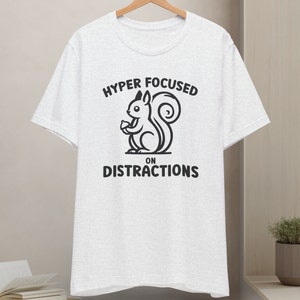 Op de afbeelding: Wit T-shirt met een zwart-witte illustratie van een eekhoorn die een eikel vasthoudt. De tekst "HYPER FOCUSED ON DISTRACTIONS" is op het T-shirt gedrukt.