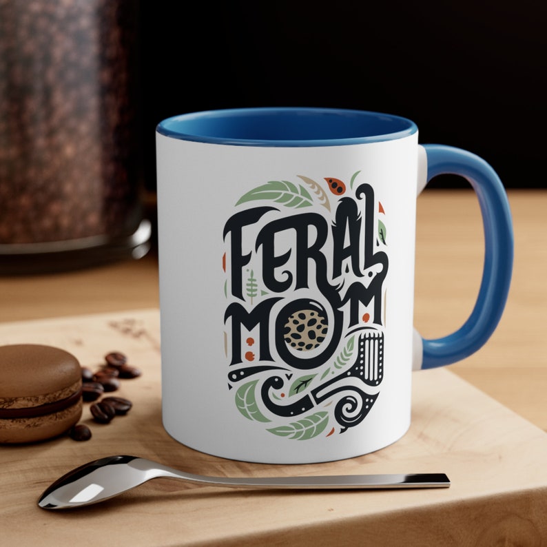 Pu&ograve; includere: Tazza in ceramica bianca con bordo e manico blu. La tazza presenta un'illustrazione in nero e verde di un motivo a foglie con il testo "Feral Mom" in lettere nere.