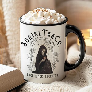 May include: White ceramic mug with a black handle and black rim. The mug has a black and white illustration of a hooded figure holding a teacup. The text on the mug reads "Suriel Tea Co. Serving Hot Tea & Not-So-Hot Loads Ever Since - Eternity".
