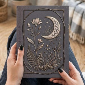 May include: A dark gray hardcover journal featuring a raised metallic bronze-toned embossed design on the front cover. The artwork depicts a crescent moon, various wildflowers, fern fronds, and small stars enclosed within a thin rectangular frame.