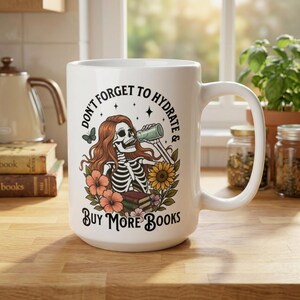 May include: White ceramic mug with a wraparound design featuring a skeleton with red hair drinking from a bottle, surrounded by flowers and the text "DON'T FORGET TO HYDRATE & BUY MORE BOOKS."