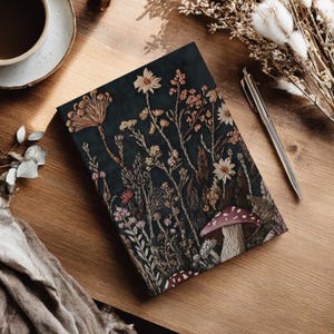 May include: A rectangular notebook featuring a dark charcoal background with a detailed botanical illustration of various wildflowers, tall grasses, and two red-capped mushrooms with white spots, rendered in muted earth tones and fine line work.