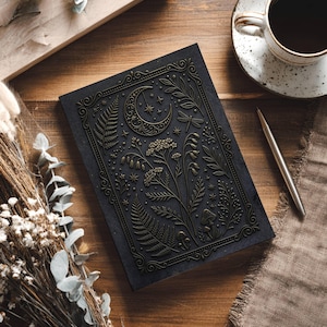 May include: A black rectangular notebook featuring an embossed gold-toned design of a crescent moon, stars, ferns, wildflowers, and mushrooms. The cover is framed by an intricate border, showcasing a detailed botanical and celestial illustration.