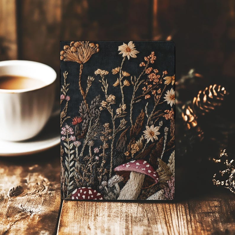 May include: A rectangular notebook featuring a dark background with intricate embroidery of various wildflowers, grasses, and two mushrooms with red caps and white spots. The design displays a variety of botanical textures and earthy tones.