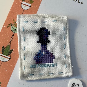 Pode incluir: Um quadrado de tecido branco com costuras azuis ao redor das bordas. O centro do quadrado apresenta um desenho de ponto cruz de uma pessoa com uma silhueta roxa e preta. O texto "remarkable" está bordado em azul abaixo da figura.