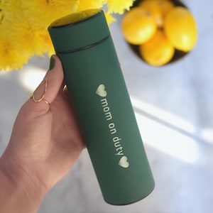 May include: A green insulated water bottle with a gold inscription that reads "mom on duty" with a heart on either side of the text.