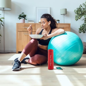 Può includere: Una donna con un reggiseno sportivo nero e leggings marroni è seduta su una palla da esercizi blu. Sta tenendo una ciotola bianca e una forchetta. Una bottiglia d'acqua rossa con il testo "Your Text Here" è sul pavimento accanto a lei.